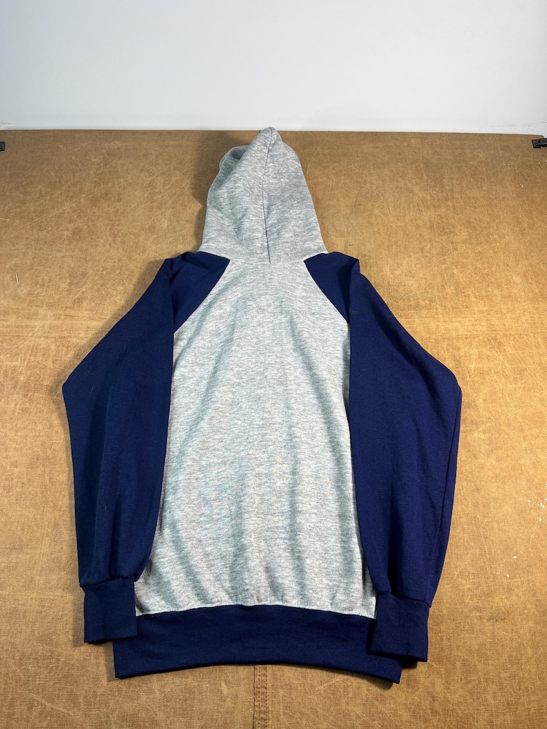 May include: A gray hooded sweatshirt with navy blue sleeves. The back of the hoodie is gray, with navy blue sleeves and cuffs. The hood is also gray. The sweatshirt is made of a soft, comfortable material.
