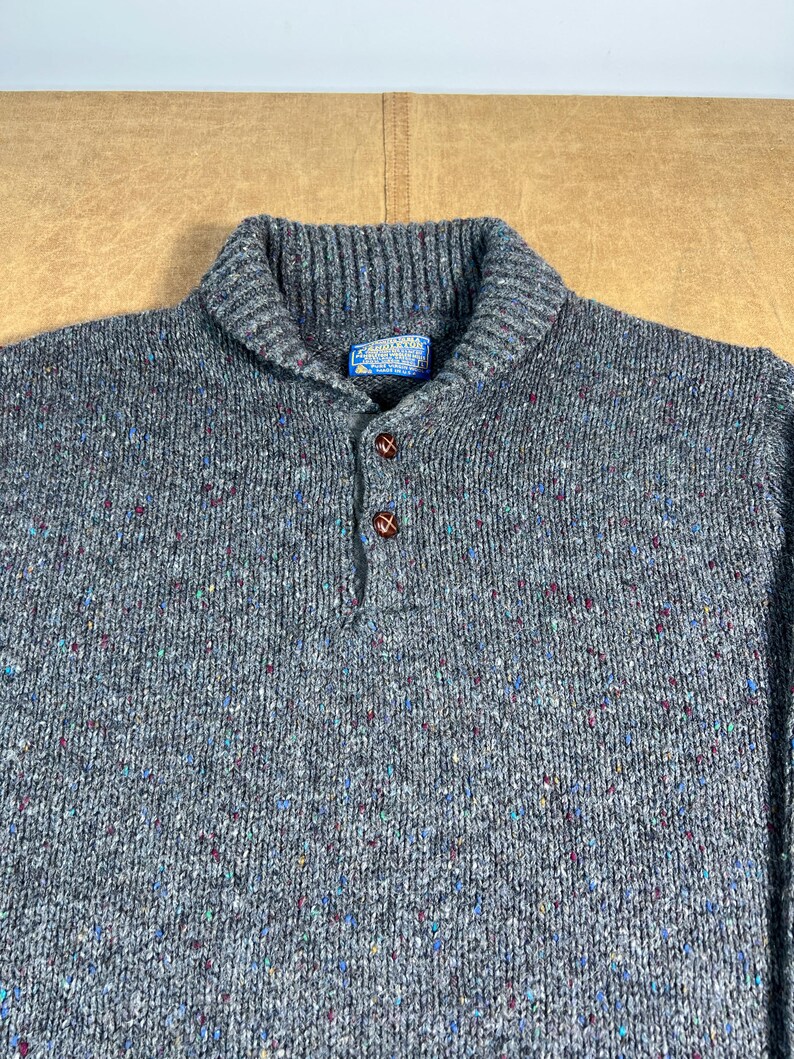May include: A gray wool sweater with a shawl collar and three brown buttons. The sweater has a speckled pattern with flecks of blue, red, and green. A small blue tag is visible near the collar.