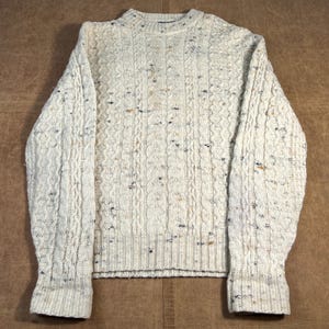 Op de afbeelding: Crèmekleurige kabelgebreide trui met spikkels van blauw, bruin en zwart. De trui heeft een ronde hals en geribde manchetten en zoom. De getextureerde knitwear is een klassieke stijl, geschikt voor koeler weer.