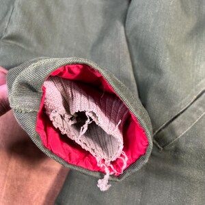 May include: Close-up of a green jacket sleeve with a red lining and a gray, ribbed cuff. The cuff is frayed and shows signs of wear. The jacket appears to be made of a durable fabric.