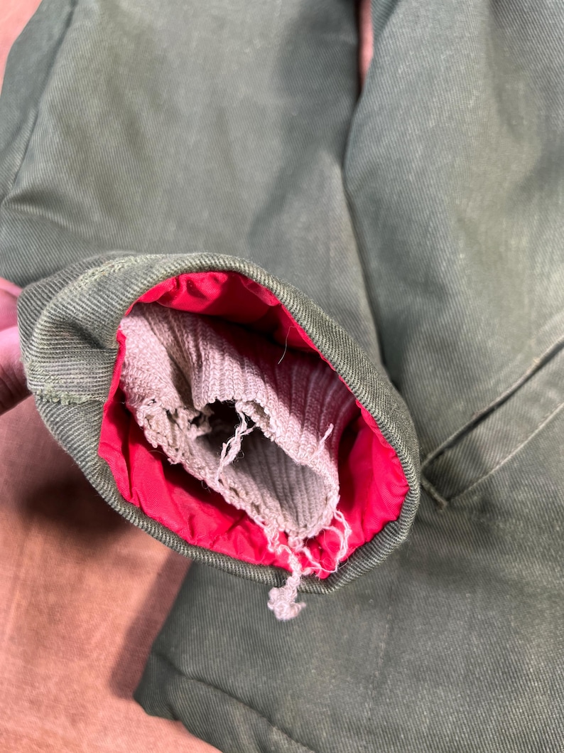 May include: Close-up of a green jacket sleeve with a red lining and a gray, ribbed cuff. The cuff is frayed and shows signs of wear. The jacket appears to be made of a durable fabric.