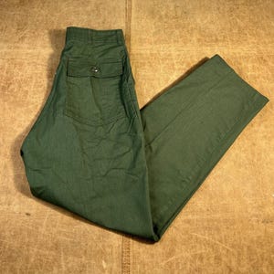 Puede incluir: Pantalones verdes oscuros con un bolsillo trasero abotonado y cierre de botón. Los pantalones están hechos de una tela resistente y se muestran sobre una superficie marrón. Diseño clásico y utilitario.