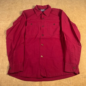 May include: A long-sleeved, claret-coloured button-down shirt with two chest pockets. The shirt features a collared neckline and buttoned cuffs. The fabric appears to be a medium-weight material. The shirt is laid flat on a neutral surface.