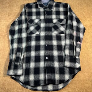May include: A black and white checked flannel shirt with two chest pockets and buttoned cuffs. The shirt features a classic collar and a curved hem. The fabric looks to be a medium-weight material, suitable for cooler weather.