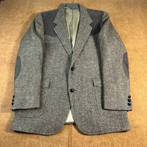 May include: A grey and white herringbone blazer featuring dark grey suede shoulder patches and matching elbow patches. The jacket has a notched lapel, two black front buttons, two flap pockets, and three black buttons on each sleeve cuff.
