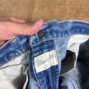 May include: Close-up of blue denim Wrangler jeans. The jeans have a button closure and a white tag with the Wrangler logo and size information. The jeans are size 34x31 and made in the USA.