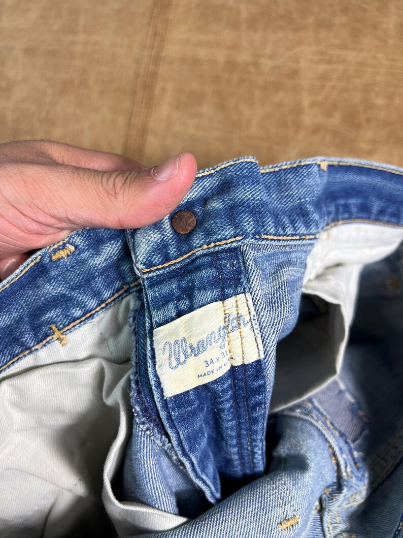 May include: Close-up of blue denim Wrangler jeans. The jeans have a button closure and a white tag with the Wrangler logo and size information. The jeans are size 34x31 and made in the USA.