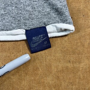 May include: Close-up of a gray t-shirt with a white trim. A navy blue tag with the Nike logo and the size "M" is attached. A white pen is in the lower left corner.