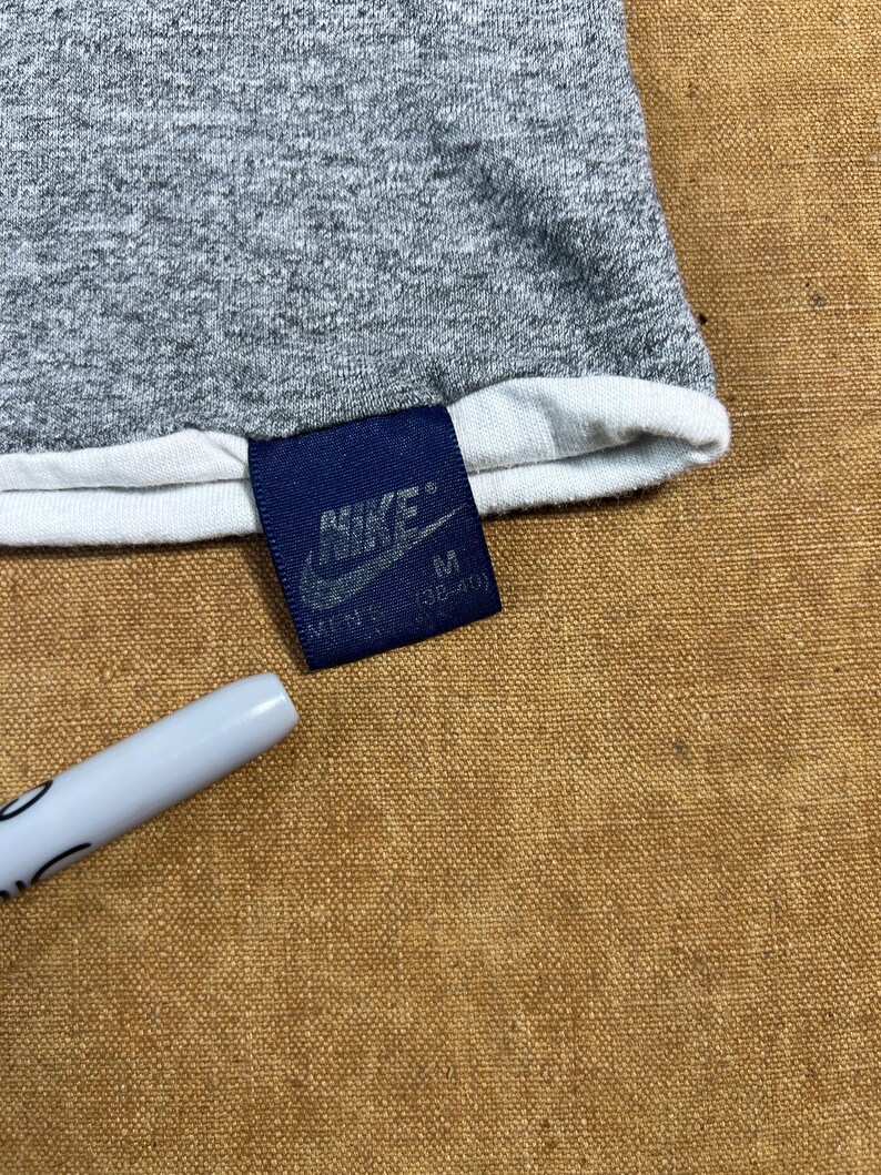 May include: Close-up of a gray t-shirt with a white trim. A navy blue tag with the Nike logo and the size "M" is attached. A white pen is in the lower left corner.