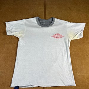 May include: White short-sleeved t-shirt with a gray crew neck and the Air Jordan logo in red on the front. The shirt has a faded appearance and a small blue tag at the bottom.