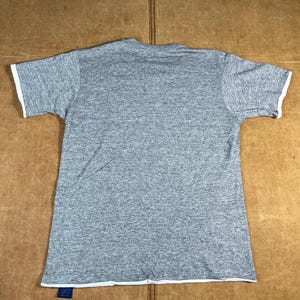 May include: A heather gray t-shirt with white trim on the sleeves and bottom hem. The short-sleeved shirt is laid flat on a brown surface. A blue tag is attached to the bottom hem.