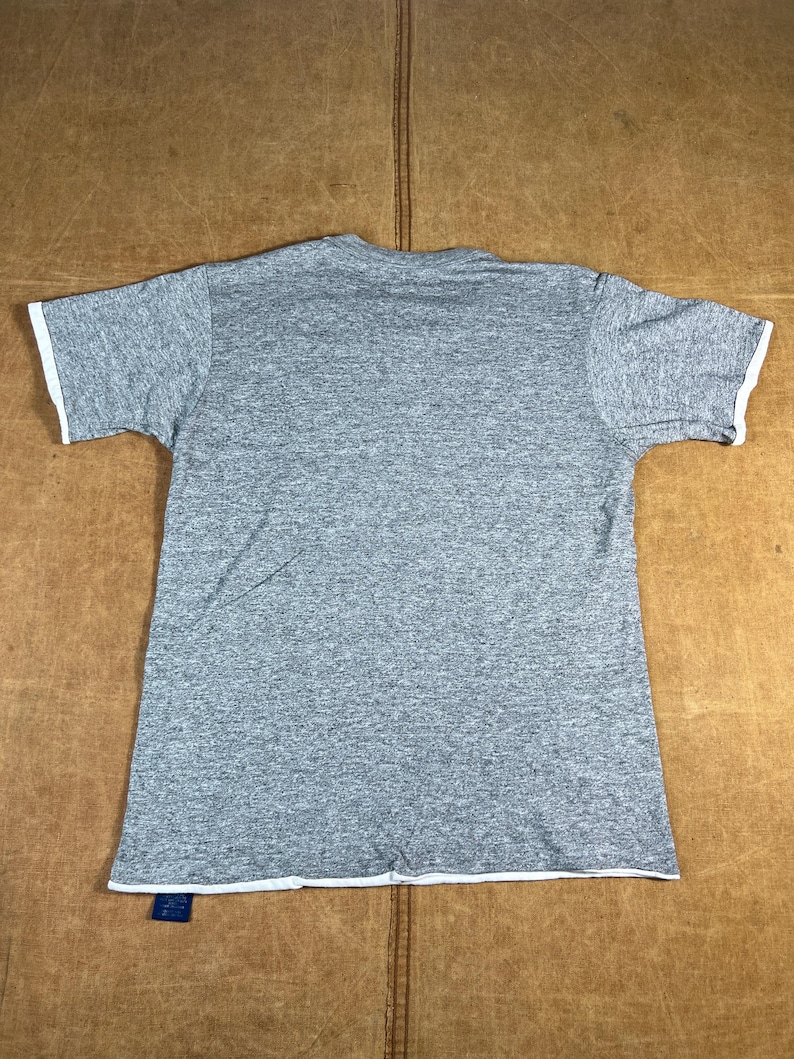 May include: A heather gray t-shirt with white trim on the sleeves and bottom hem. The short-sleeved shirt is laid flat on a brown surface. A blue tag is attached to the bottom hem.