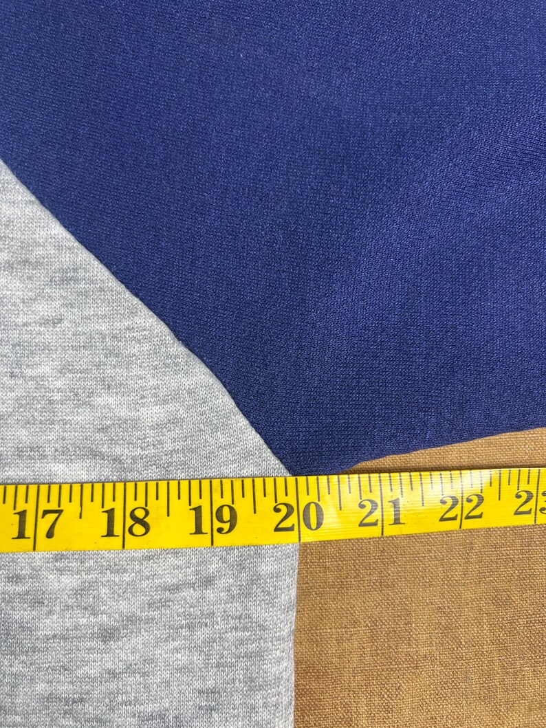 May include: Close-up of a blue and gray raglan sleeve shirt. A yellow measuring tape is laid across the shirt, showing measurements from 17 to 23 inches.
