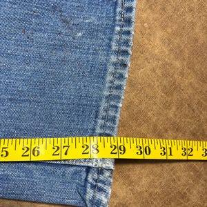 May include: Close-up of light blue denim jeans with a yellow measuring tape showing a waist size of approximately 29 inches. The jeans have brown spots and a visible seam.
