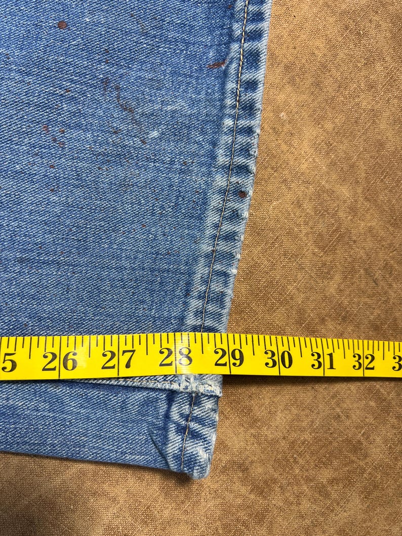 May include: Close-up of light blue denim jeans with a yellow measuring tape showing a waist size of approximately 29 inches. The jeans have brown spots and a visible seam.