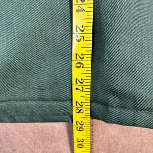 May include: A close-up shot of a green fabric with a yellow measuring tape. The tape shows measurements from 23 to 32 inches. The fabric appears to be a solid color, possibly a garment or textile.