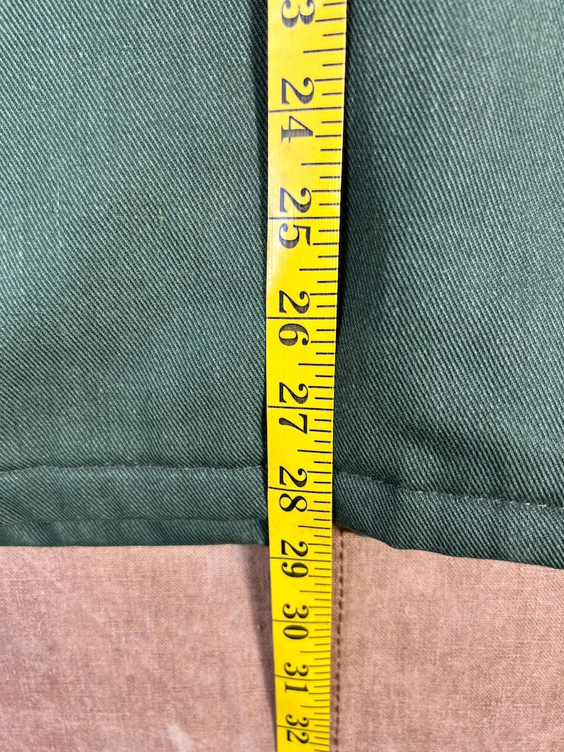 May include: A close-up shot of a green fabric with a yellow measuring tape. The tape shows measurements from 23 to 32 inches. The fabric appears to be a solid color, possibly a garment or textile.