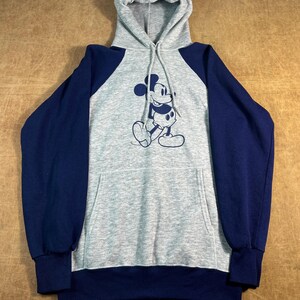 May include: A gray hooded sweatshirt with navy blue sleeves and a front pocket. The sweatshirt features a navy blue graphic of Mickey Mouse on the front. The hood and body are gray.