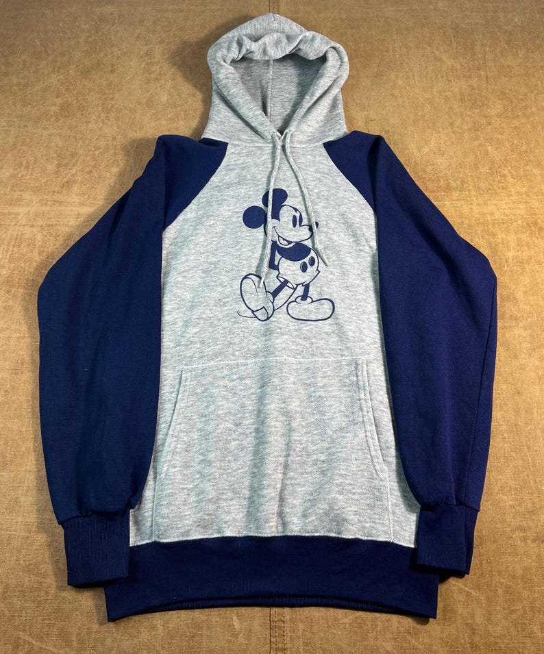 May include: A gray hooded sweatshirt with navy blue sleeves and a front pocket. The sweatshirt features a navy blue graphic of Mickey Mouse on the front. The hood and body are gray.
