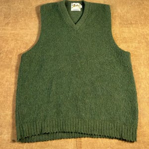 Puede incluir: Un chaleco de punto verde con cuello en V de lana. El chaleco tiene un dobladillo inferior acanalado y una pequeña etiqueta en la parte posterior del cuello. El chaleco es de color verde liso y está hecho de un material suave y texturizado.