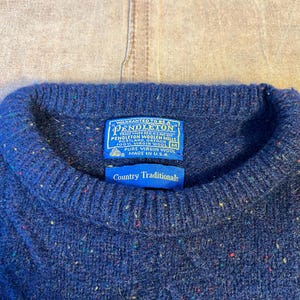 May include: Close-up of a blue Pendleton wool sweater with a crew neck. The sweater has flecks of color throughout and a blue tag that reads "Country Traditionals" and "100% Virgin Wool". The tag also states "Made in U.S.A."
