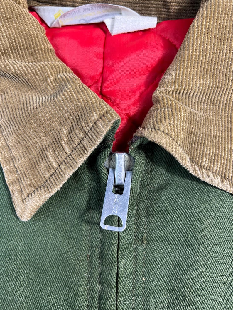 May include: Close-up of a green jacket with a brown corduroy collar and a red quilted lining. The jacket has a silver zipper. The label inside the collar reads "MACHINE WASH".