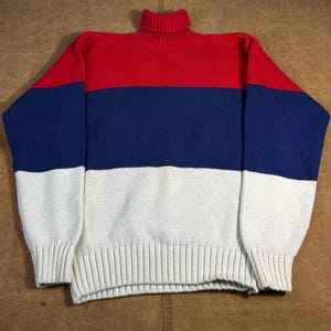 May include: A color-blocked turtleneck sweater in red, navy blue, and white. The sweater has a ribbed collar, cuffs, and hem. The sleeves are long, and the body is a relaxed fit. The sweater is made of a soft, knitted material.