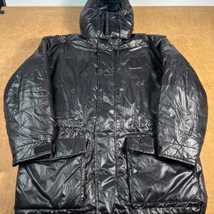 Könnte beinhalten: Ein schwarzer Wintermantel mit Kapuze und glänzender, gesteppter Optik. Der Mantel hat eine Kapuze mit Kordelzug, mehrere Taschen und eine verstellbare Taille. Der Markenname ist auf der Brust sichtbar. Für kaltes Wetter konzipiert.