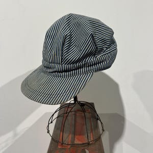 May include: A railroad-style engineer cap featuring a classic hickory stripe pattern in dark navy blue and off-white. The cap has a rounded crown with a gathered fabric band and a stiff, curved brim, showcasing a traditional workwear aesthetic.