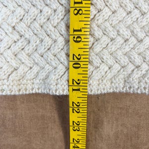 May include: Close-up of a cream-colored knitted fabric with a textured, woven pattern. A yellow measuring tape is positioned vertically, showing measurements from 17 to 25 inches. The fabric's texture is visible.