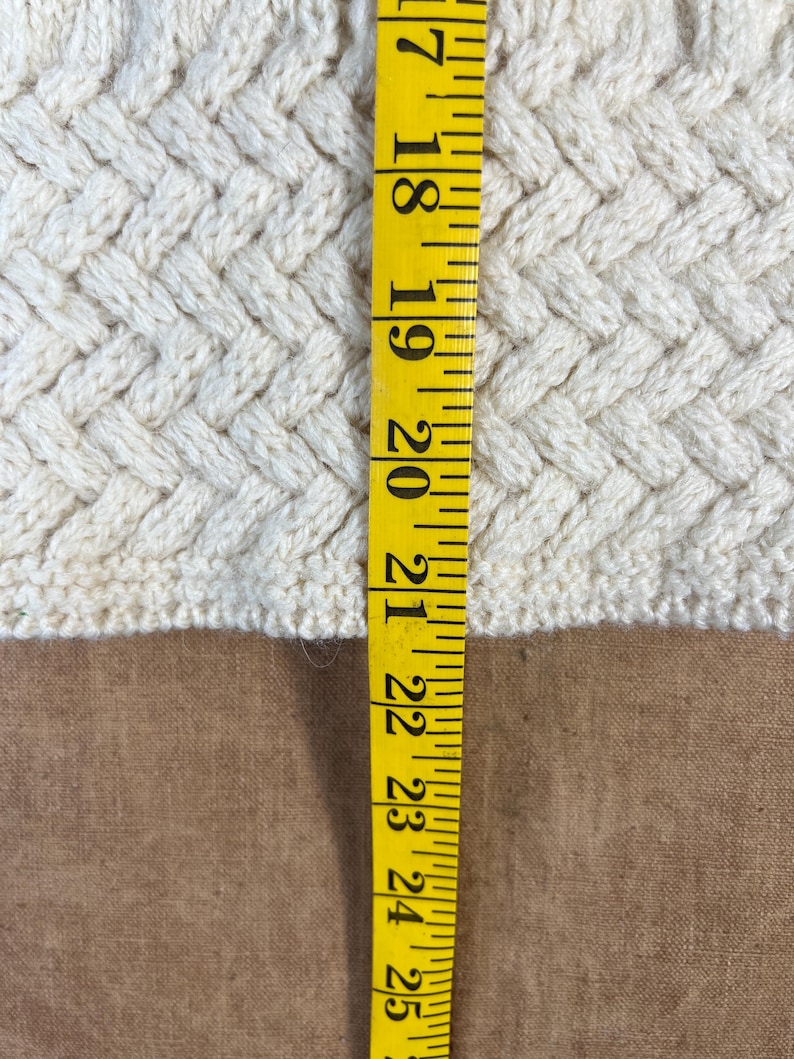 May include: Close-up of a cream-colored knitted fabric with a textured, woven pattern. A yellow measuring tape is positioned vertically, showing measurements from 17 to 25 inches. The fabric's texture is visible.