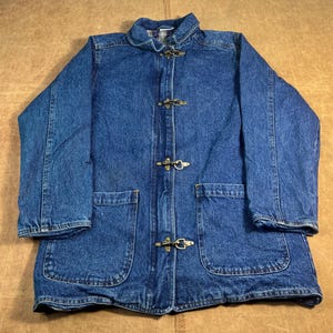 May include: A blue denim jacket with a collared neckline and three toggle closures. The jacket has two large front pockets and a plaid lining visible at the collar. The jacket is made of a medium-wash denim material.