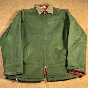 May include: An olive green work jacket with a tan corduroy collar and a red interior lining. The jacket has a zip closure, two front pockets, and a small chest pocket. The jacket is a vintage style.