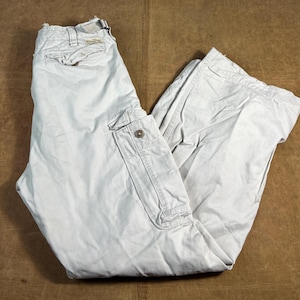 May include: Light beige cargo trousers with multiple pockets and a button closure. The trousers are made of a durable fabric and have a relaxed fit. The trousers are folded on a brown surface.