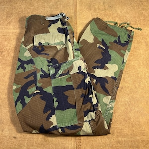 Peut inclure: Pantalon cargo camouflage avec un motif forestier, composé de nuances de vert, de marron et de noir. Le pantalon comporte plusieurs poches, une fermeture à bouton et à glissière, et une boucle de ceinture. Le tissu semble être un matériau durable de style militaire.