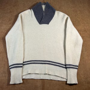 May include: Cream-colored waffle knit sweater with a gray shawl collar and cuffs. The sweater features two gray stripes around the waist and cuffs. The sweater is made of a textured knit fabric.