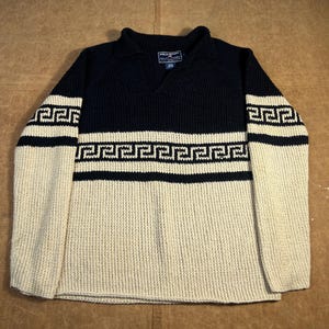 Peut inclure: Un pull bleu marine et crème avec un motif géométrique. Le pull a un col en V et des manches longues. Les manches et le bas du corps sont de couleur crème, tandis que le haut du corps est bleu marine. Le pull a un motif géométrique sur la poitrine et les manches.