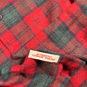May include: A close-up shot of a red and green tartan fabric. The plaid design has a traditional pattern of intersecting lines. A small white label reads "AUTHENTIC LINDSAY TARTAN". The material looks like a woven fabric, possibly wool or a wool mix.