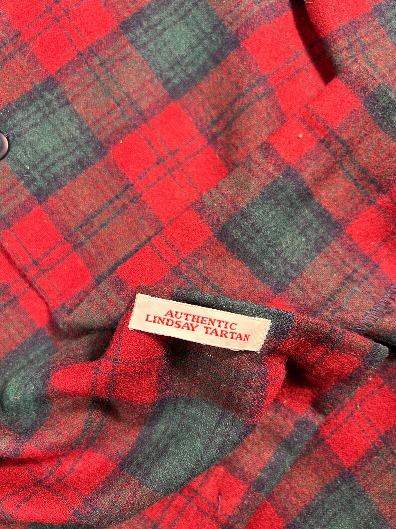 May include: A close-up shot of a red and green tartan fabric. The plaid design has a traditional pattern of intersecting lines. A small white label reads "AUTHENTIC LINDSAY TARTAN". The material looks like a woven fabric, possibly wool or a wool mix.