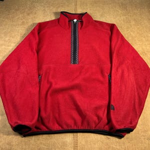 May include: A red fleece pullover with a quarter-zip closure and black trim. The pullover has a high collar and a small pocket on the left chest. The North Face logo is visible on the lower left side. The fleece is a warm, casual garment.