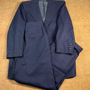 Op de afbeelding: Marineblauw krijtstreep pak. De set bevat een blazer met drie knopen op elke manchet en een bijpassende broek. De binnenvoering van de blazer heeft een ruitpatroon.