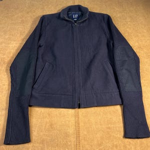 May include: A navy blue Gap jacket with a ribbed collar and cuffs. The jacket features a full zipper, two front pockets, and black patches on the elbows. The Gap logo is visible on the collar.