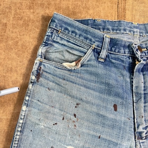 May include: Close-up of a pair of worn blue denim jeans. The jeans show signs of wear and tear, with faded areas and brown paint splatters. The pocket lining is visible, and the jeans are on a brown surface.