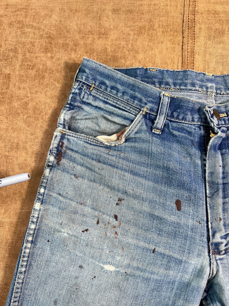 May include: Close-up of a pair of worn blue denim jeans. The jeans show signs of wear and tear, with faded areas and brown paint splatters. The pocket lining is visible, and the jeans are on a brown surface.
