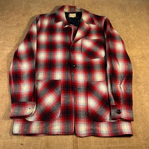 Op de afbeelding: Een rood, zwart en crèmekleurig geruit jack met een kraag, twee grote voorzakken en knoopsluitingen. De jas heeft een klassieke, vintage stijl.
