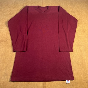 May include: A long-sleeved, burgundy-colored shirt with a V-neck and reinforced shoulder panels. The shirt has a relaxed fit and a tag at the bottom hem. The fabric appears to be a soft, knit material.