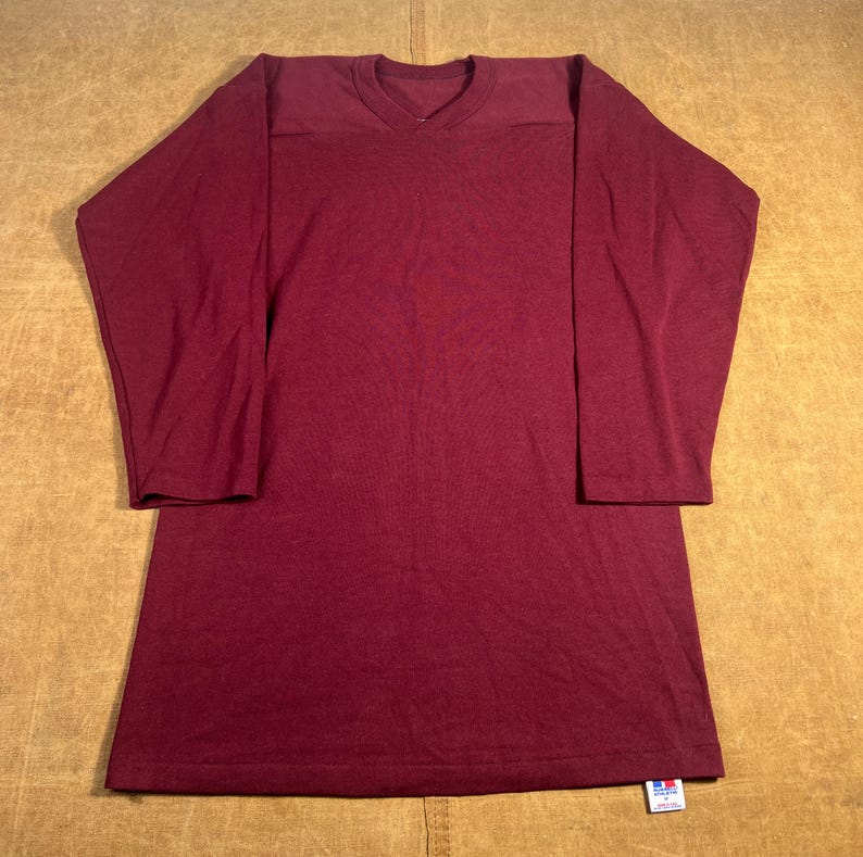 May include: A long-sleeved, burgundy-colored shirt with a V-neck and reinforced shoulder panels. The shirt has a relaxed fit and a tag at the bottom hem. The fabric appears to be a soft, knit material.