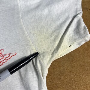 May include: Close-up of a white t-shirt with a red graphic design on the front. The shirt has short sleeves and appears to be made of a lightweight fabric. A black marker is placed on the shirt.
