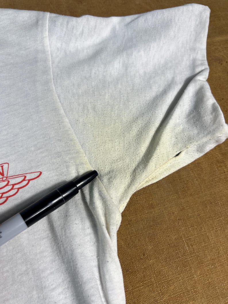 May include: Close-up of a white t-shirt with a red graphic design on the front. The shirt has short sleeves and appears to be made of a lightweight fabric. A black marker is placed on the shirt.