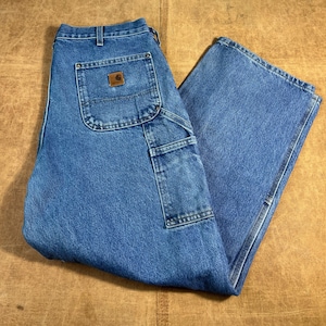 May include: Blue denim work trousers with multiple pockets and a brown leather patch on the back pocket. The trousers have a relaxed fit and are made of durable denim material. The Carhartt logo is visible on the back pocket.